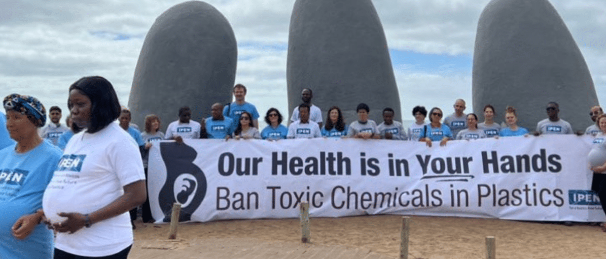 IPEN members holding a banner saying "Our Health is in Your Hands" in front of a sculpture of fingers coming up from the sand