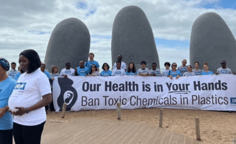IPEN members holding a banner saying "Our Health is in Your Hands" in front of a sculpture of fingers coming up from the sand