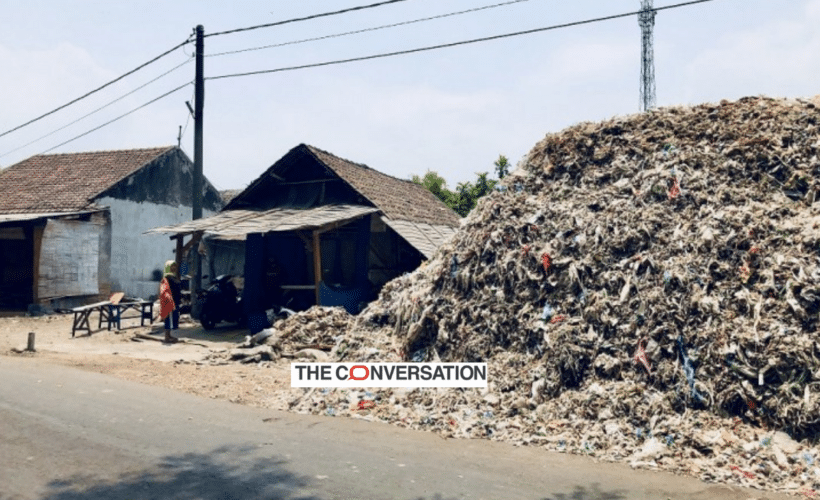 a huge pile of plastic waste next to a house, logo of The Conversation in foreground