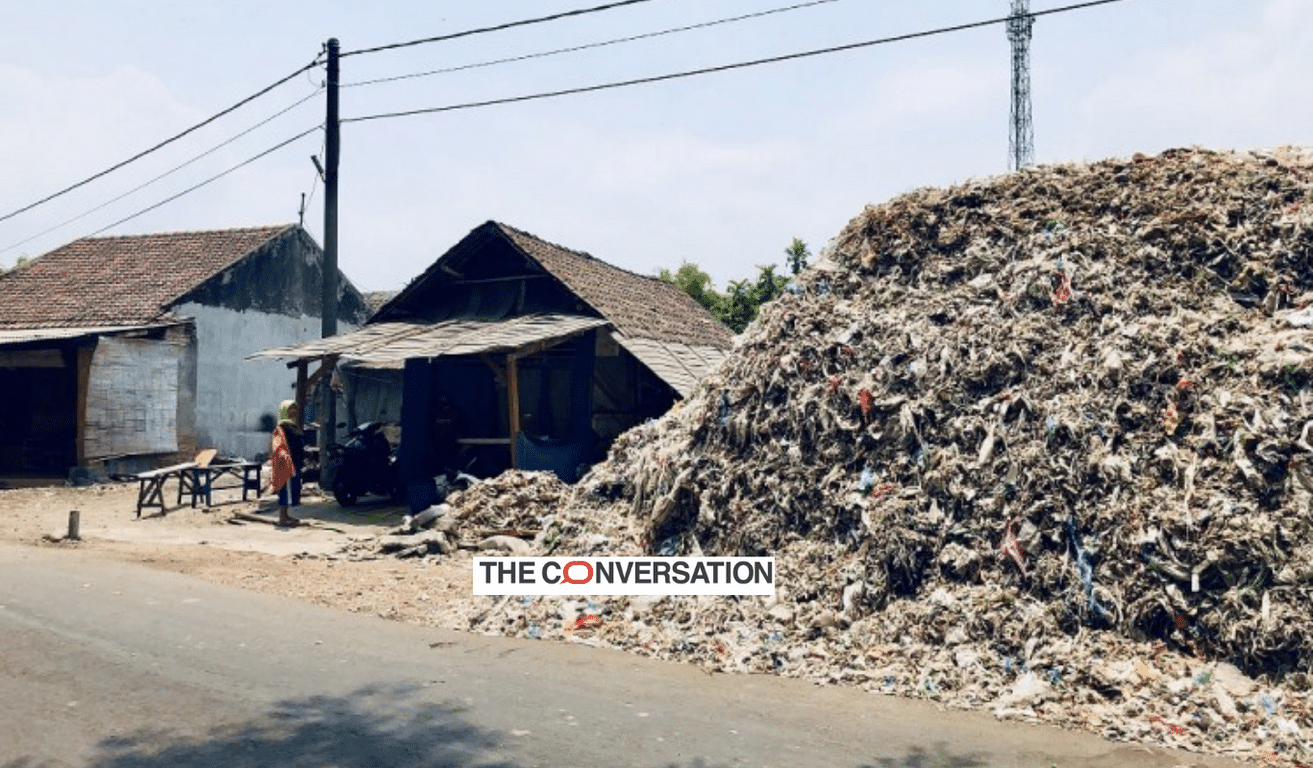 a huge pile of plastic waste next to a house, logo of The Conversation in foreground